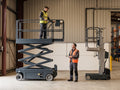 A learner on a scissor lift platform beside a vertical personnel lift, with an instructor on the floor