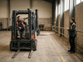 A learner seated on a parked forklift while an instructor speaks from a separate pedestrian walkway